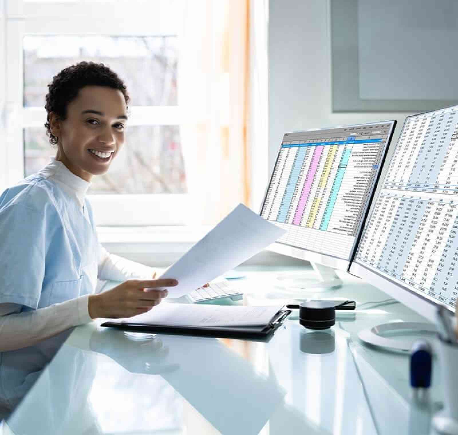 A professional medical billing specialist sits at a desk working with paperwork and dual computer monitors displaying spreadsheets and billing data, representing a role in medical billing and revenue cycle management.